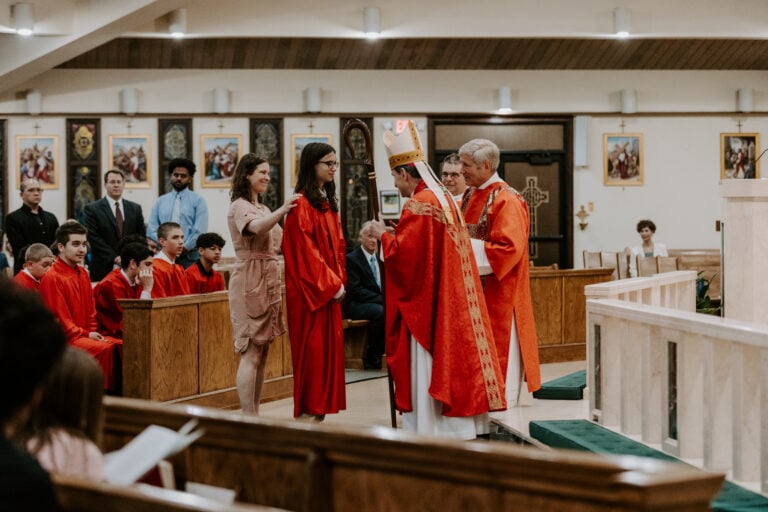confirmation ceremony at a church with a bishop in red vestments, a young woman in red robes being confirmed, and a woman standing next to her. several boys in red robes are seated in the pews, and other attendees are visible in the background.